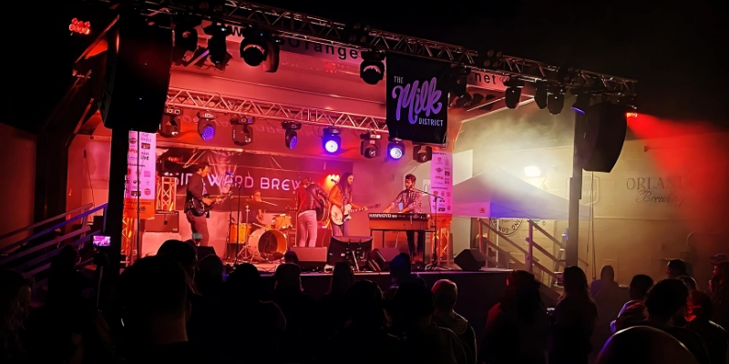 A lively band takes the stage at night, surrounded by striking red lights, captivating the audience in Downtown Orlando's Milk District.