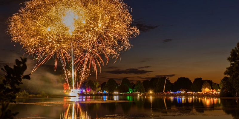 Vibrant fireworks burst above a serene lake at night, surrounded by trees, as part of the Downtown Orlando Events festivities.
