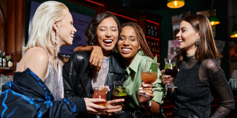 Four women happily pose with drinks at a bar, enjoying an Orlando bachelorette party-themed bar crawl.