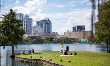 A group enjoys the grass by a lake, with the skyline of Downtown Orlando rising in the background, highlighting city life.